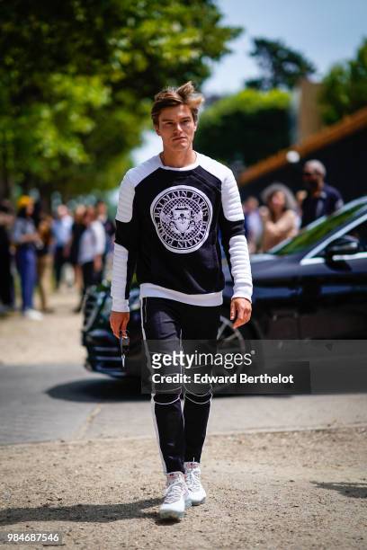 Oliver Cheshire wears a Balmain black and white pull over, black pants, white sneakers, outside Balmain, during Paris Fashion Week - Menswear...