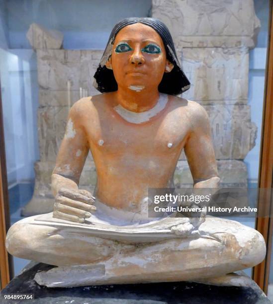 Painted limestone statue of an Egyptian scribe seated cross-legged holding an open papyrus scroll. 5th Dynasty. Saqqara.