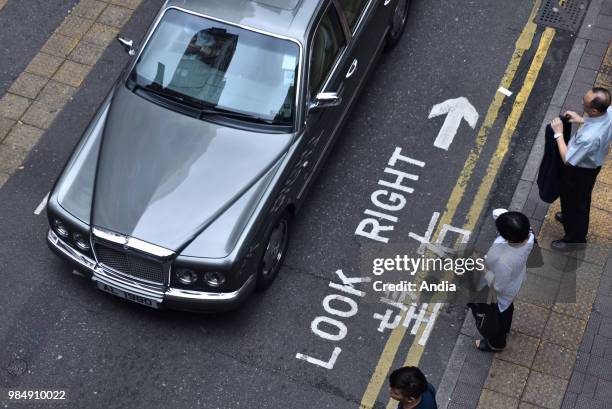 Hong Kong. Pedestrian waiting on a pavement, with road marking in English and Chinese: 'Look right, look left', and looking at a luxury car to...