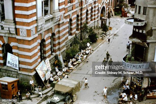Elevated view of a street vendors outside the 'Hotel Namsan' and a 'Portrait Studio' , while a covered military jeep drives one direction on the...
