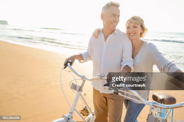 coppia matura in bicicletta sulla spiaggia al tramonto o all'alba - mature couple foto e immagini stock