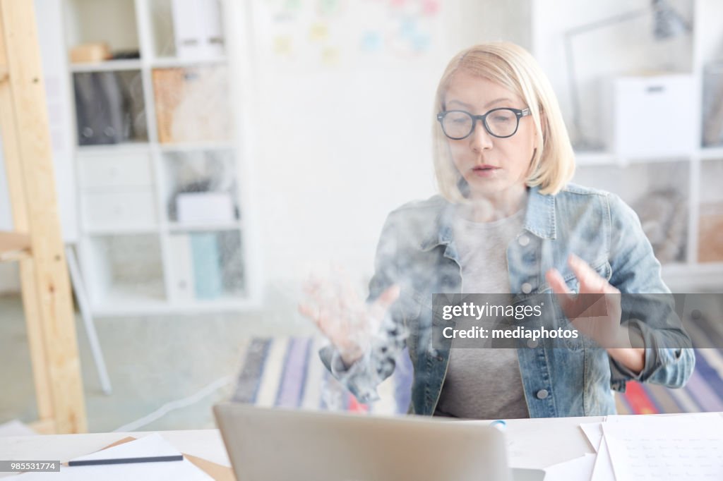 Scared middle-aged freelance woman waving hands and blowing at smoking laptop while working at home