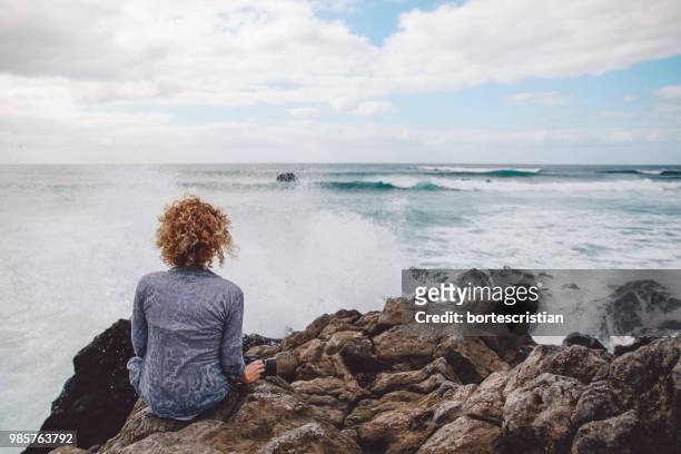 rear view of woman sitting on rock by sea against sky - bortes photos et images de collection