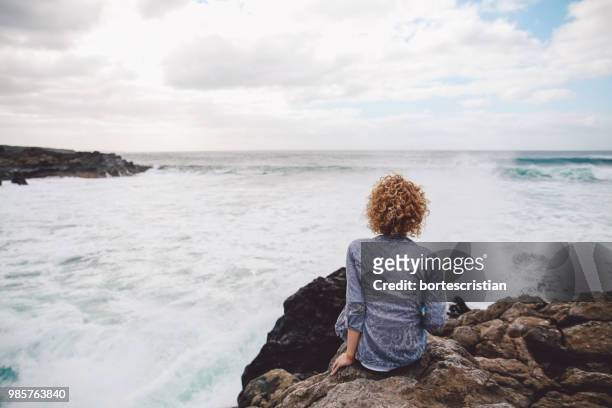 rear view of woman sitting on rock by sea against sky - bortes photos et images de collection