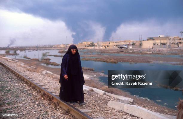 Shi'a woman walks by the railroad track on the outskirts of Baghdad with a huge smoke blanket covering the skyline of the city, during the Gulf War,...