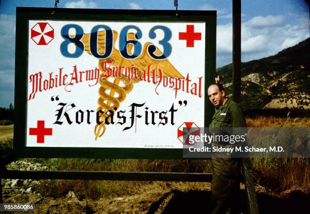 Portrait of American military surgeon Captain Sidney Schaer as he poses with the large sign for his unit, the 8063rd MASH , which also reads 'Korea's...