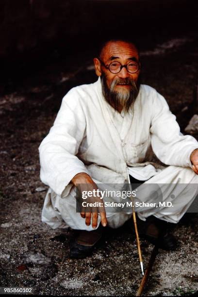 Portrait of an unidentified, bearded man dressed in white as he crouches down, a long pipe in his lap, South Korea, January 1952.