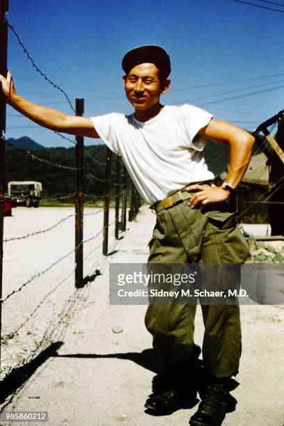 Portrait of an unidentified man dressed in a t-shirt and wearing military-issue trousers as he leans on a barbed wire fence at the 8063rd MASH ,...