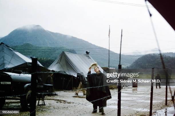 View of Captain Jim C Jones, of the 8063rd MASH , dressed in a poncho, as he doffs a straw hat in the rain, South Korea, January 1952.