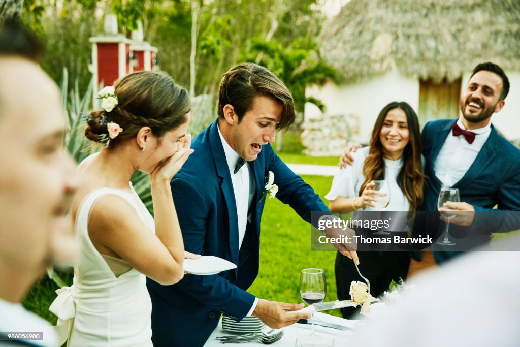 Laughing bride and groom cutting cake for guests during outdoor wedding reception