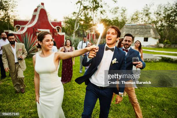 Bride and groom holding sparkler while celebrating during outdoor wedding reception with friends