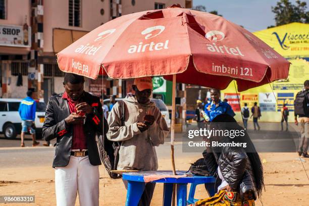 Pedestrians check their mobile phones under the shelter of parasol featuring a Bharti Airtel Ltd. Logo in Lilongwe, Malawi, on Tuesday, June 26 2018....