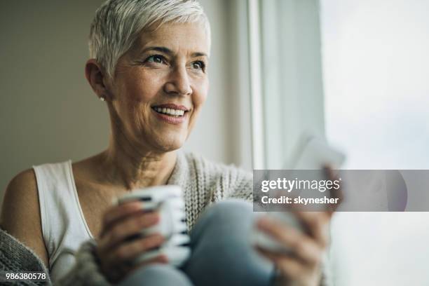 happy mature woman using mobile phone by the window. - 2017 common good forum stock pictures, royalty-free photos & images