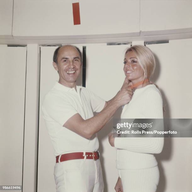 French fashion designer Andre Courreges pictured dressed all in white as he ties a bow around the next of a female fashion model in 1970.