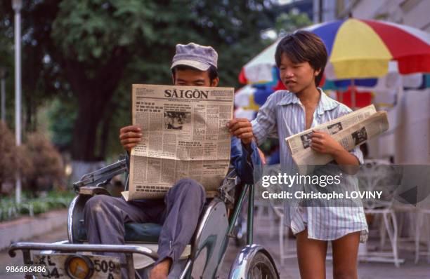 Une homme lit le journal accompagné d'un jeune garçon dans une rue de Hô-Chi-Minh-Ville en octobre 1989, Viet Nam.