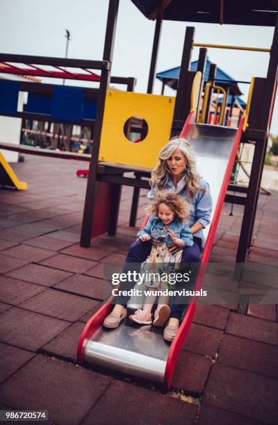 fashionable grandmother and little granddaughter sliding down playground slide - great granddaughter stock pictures, royalty-free photos & images
