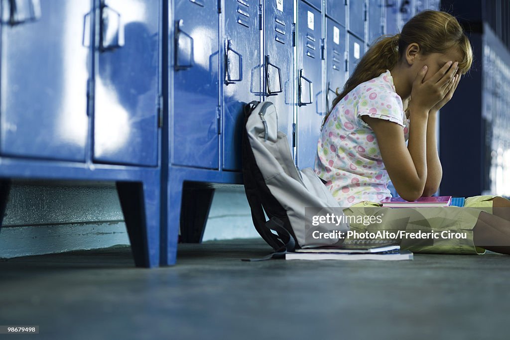 Female junior high student sitting on hall floor near lockers, upset and covering face with hands