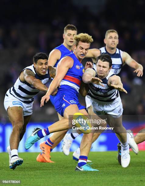 Mitch Wallis of the Bulldogs kicks whilst being tackled by Tim Kelly and Patrick Dangerfield of the Cats during the round 15 AFL match between the...