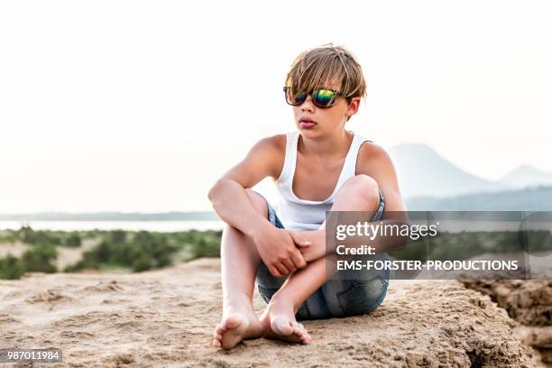 bored preadolescent boy with long blonde hair sits on stone cliff near the beach - ems forster productions 個照片及圖片檔