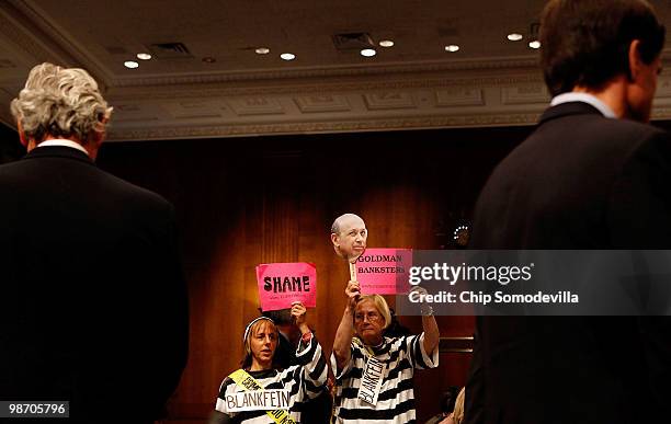 Demonstrators from Code Pink for Peace hold signs behind executives from the The Goldman Sachs Group during a break in a hearing of the Senate...