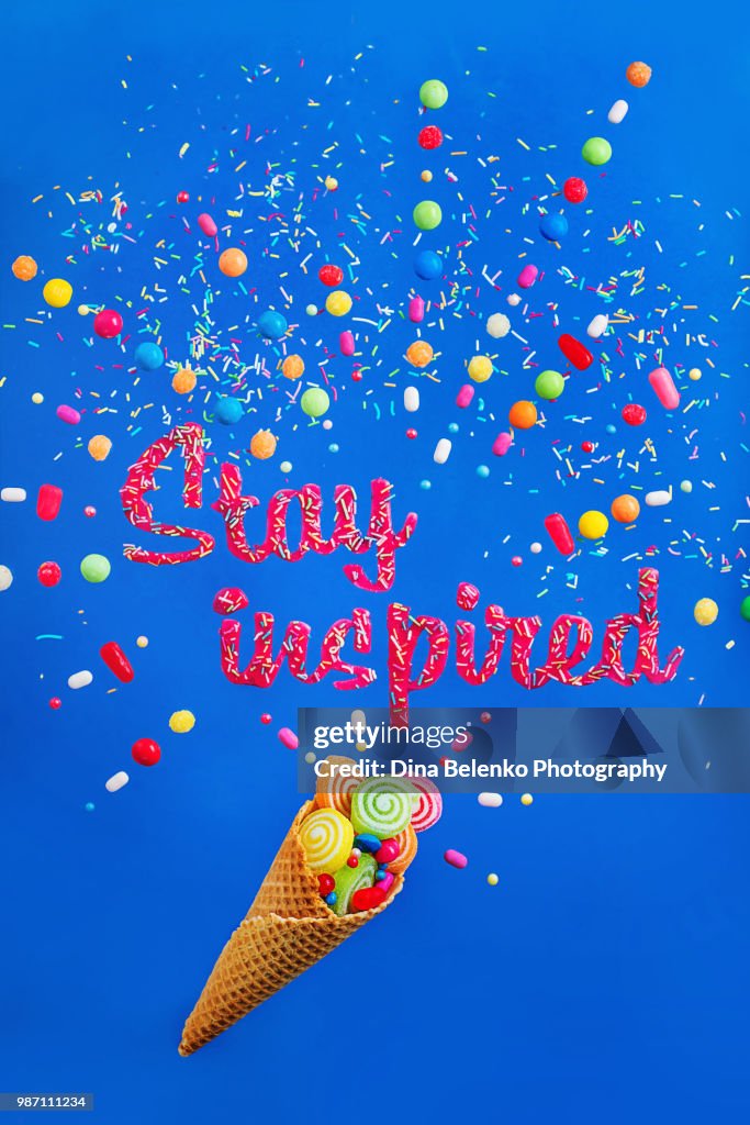 Ice cream waffle cone on a vibrant background with words Stay inspired made with glazing and sprinkles. Sweet motivation flat lay with candies and bonbons.