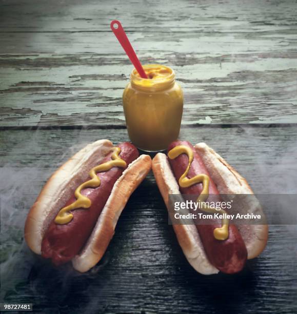 Close-up of two steaming hot dogs on a wooden table, with a jar of mustard, 1980.