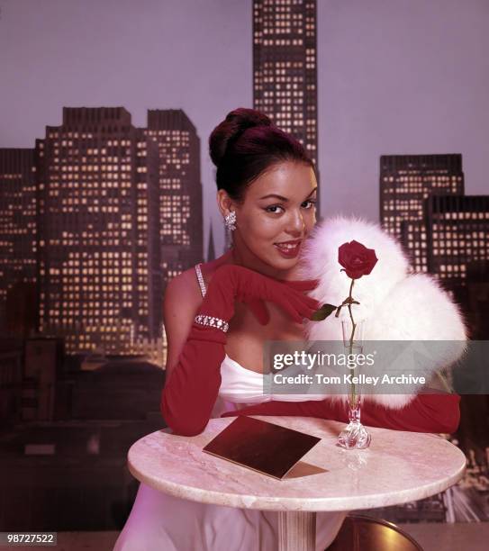 Studio portrait of an unidentified model in a white evening gown with a fur stole and red elbow gloves as she sits at a small, round marble table in...