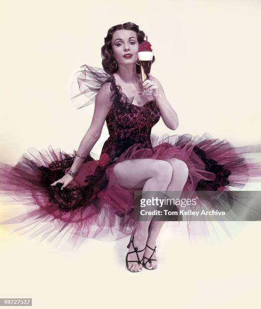 Studio portrait of an unidentified model, in a red and black cocktail dress with a chiffon skirt, as she poses with a glass of beer in one hand, 1953.