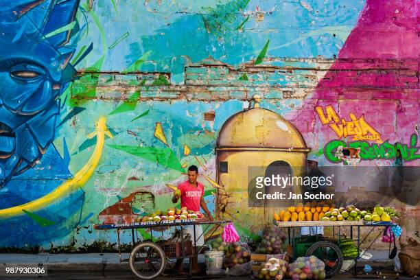 Colombian vendors sell fruits in front of a large mural in Getsemaní, a popular artistic neighborhood on December 12, 2017 in Cartagena, Colombia....