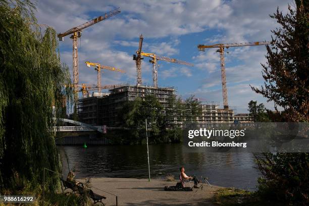 Construction cranes stand above the Europacity project residential building site as a fisherman sits beside his rod on the bank of the River Spree in...