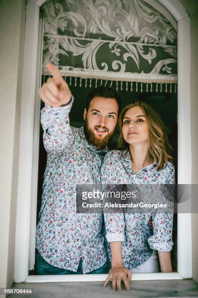 young couple in same shirts looking out the window of their apartment - cool couple in apartment stock pictures, royalty-free photos & images