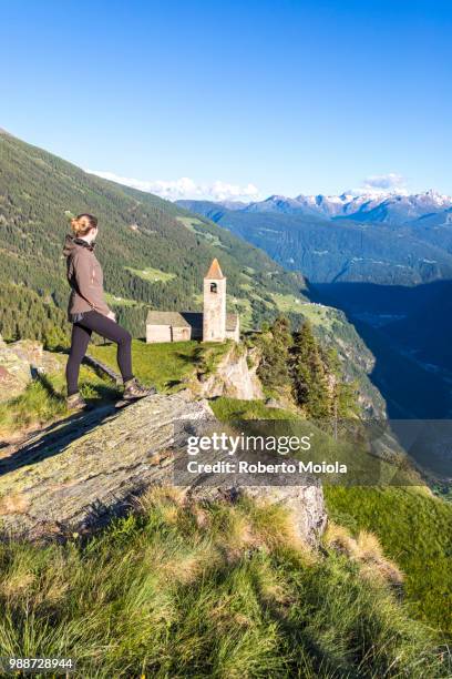 woman looks at old church perched on mountains, san romerio alp, brusio, canton of graubunden, poschiavo valley, switzerland, europe - graubunden canton fotografías e imágenes de stock