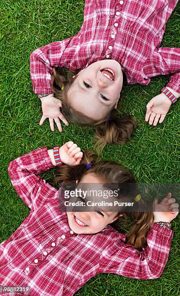 twin girls lying on grass looking up and smiling - newpremiumuk stock-fotos und bilder