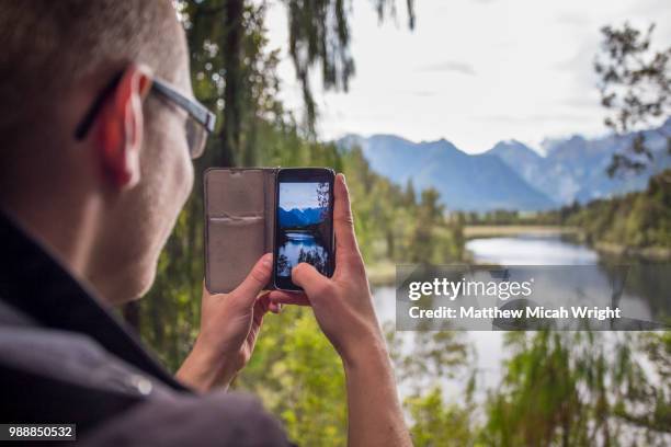 hikers explore the lake matheson trails. the lakes mirror reflections are a famous photo stop for visitors as a man take a photo with his phone. - south westland stock-fotos und bilder