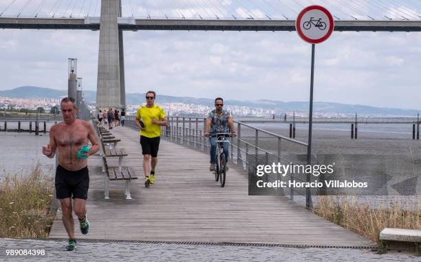 Joggers run while a cyclist rides on the North end of Parque das Nacoes , in front of Vasco da Gama bridge during a sunny Sunday morning on July 01,...