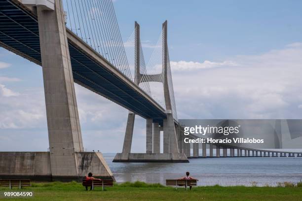 Two women sit on benches by the Vasco da Gama bridge during a sunny Sunday morning on July 01, 2018 in Lisbon, Portugal. Sunny weather is one of the...