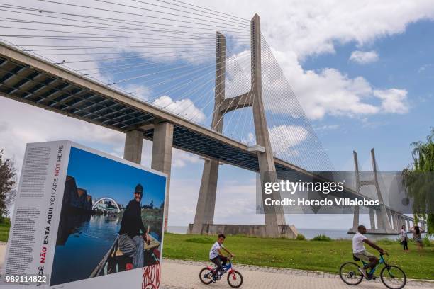 Man and a child cycle near Vasco da Gama bridge by the open air photo exhibition "Voce Nao Esta Aqui" by Portuguese photojournalist Bruno Portela,...