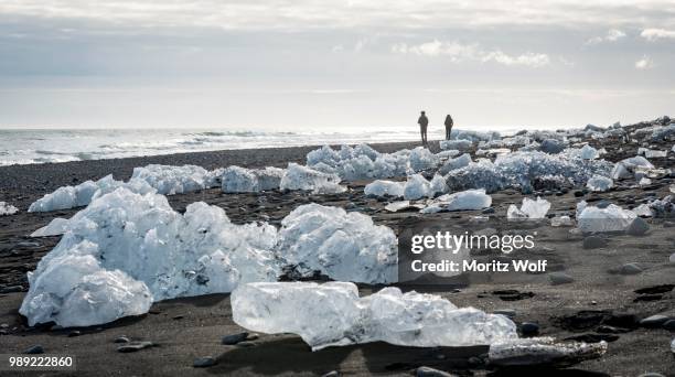 two tourists walking on the beach, ice floes on black lava sand, diamond beach, fjallsarlon glacier lagoon, east iceland, iceland - austurland stock pictures, royalty-free photos & images