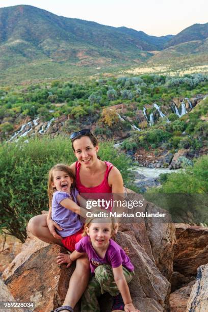 mother with two girls sitting on rocks in green landscape, behind epupa falls, kunene, kunene region, namibia - kunene region stock pictures, royalty-free photos & images