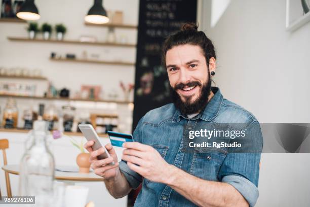 smiling young man holding up a credit card and a mobile phone - earring card stock pictures, royalty-free photos & images