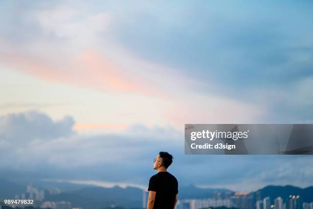 young man enjoying the tranquility while gazing at dramatic sky in deep thought - mirando al cielo fotografías e imágenes de stock