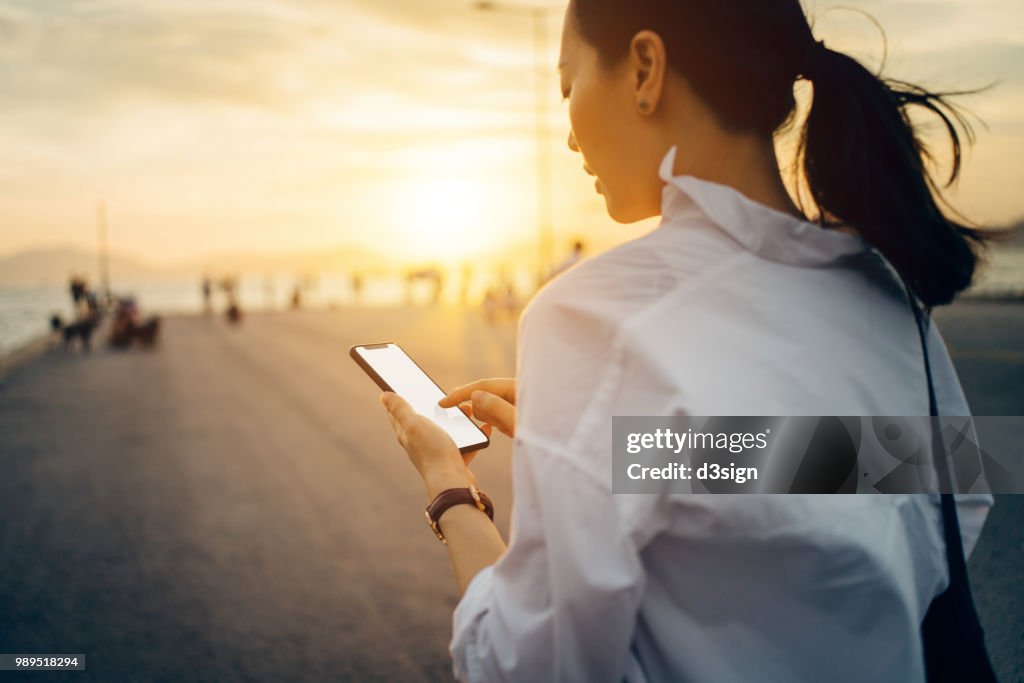 Young lady using smartphone while having a relaxing walk by the pier afterwork at sunset
