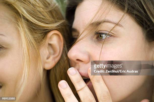 young woman whispering secret into friend's ear, close-up - confidentiality stockfoto's en -beelden