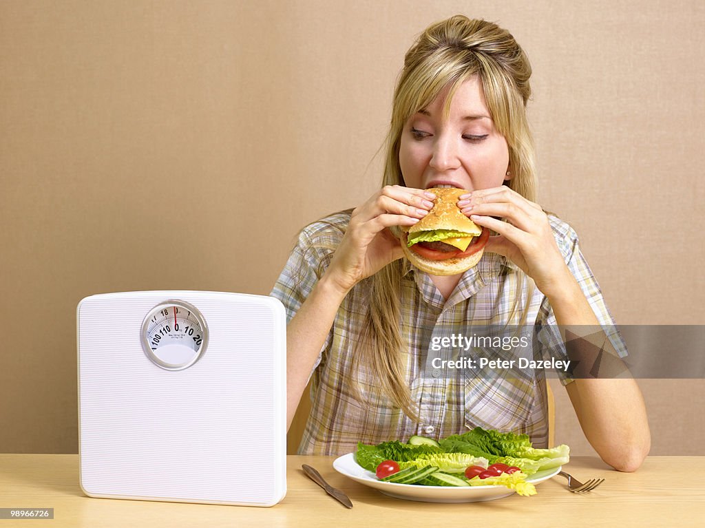 Bulimic girl eating burger with bathroom scales