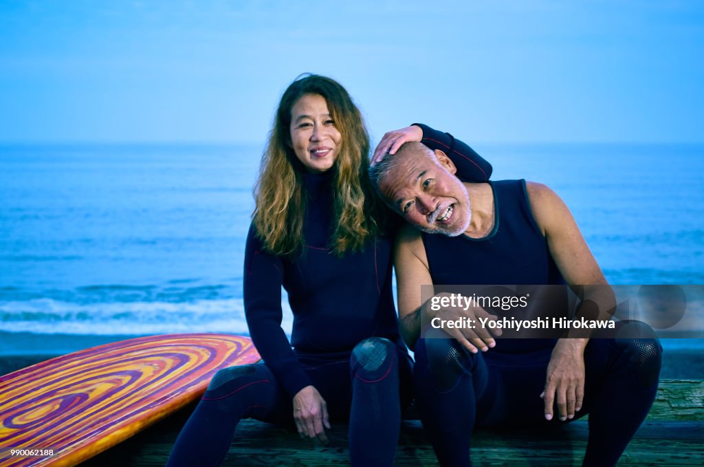 Senior couple sit on the sea sand before surfing early in the morning next to surfboard