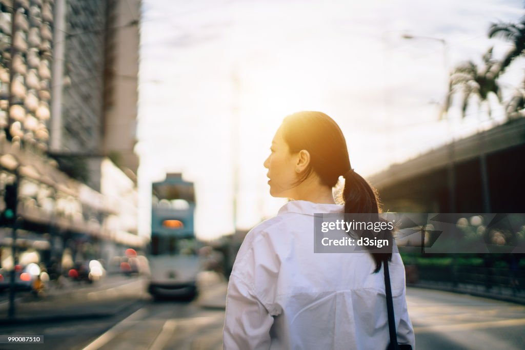 Beautiful young woman waiting for transportation ride on city street at sunset