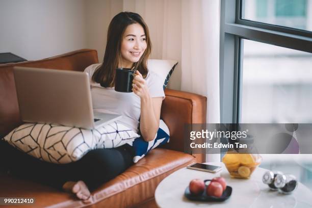een aziatische chinese vrouw zittend op de bank in de woonkamer kijken laptop haar mobiel apparaat houden van een kopje koffie - asian cup stockfoto's en -beelden