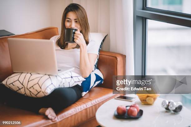 een aziatische chinese vrouw zittend op de bank in de woonkamer kijken laptop houden van een kopje koffie - asian cup stockfoto's en -beelden