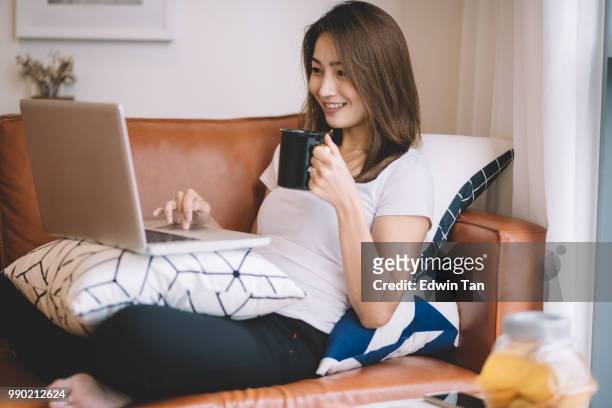 een aziatische chinese vrouw zittend op de bank in de woonkamer kijken laptop haar mobiel apparaat houden van een kopje koffie - asian cup stockfoto's en -beelden
