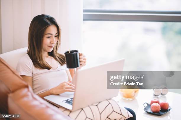 een aziatische chinese vrouw zittend op de bank in de woonkamer kijken laptop haar mobiel apparaat houden van een kopje koffie - asian cup stockfoto's en -beelden